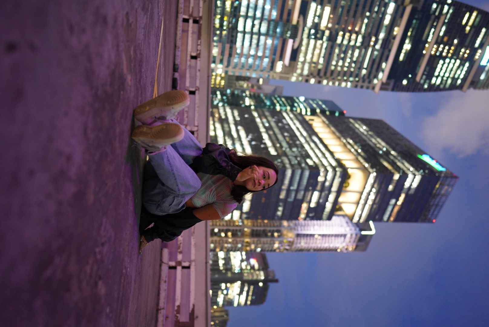 A young woman sitting on a rooftop parking lot at night with a city skyline with illuminated tall buildings in the background.